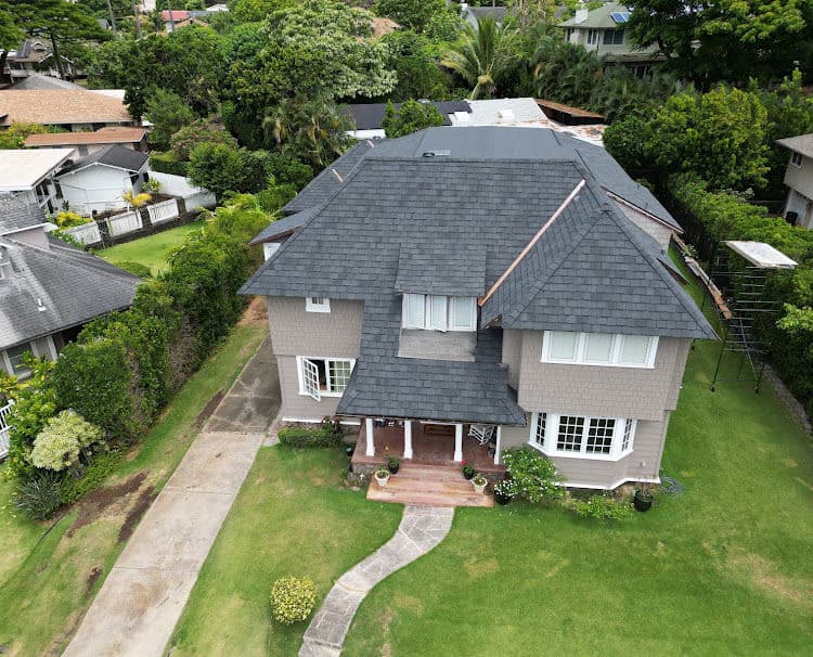 Aerial view of a large gray house with a lush green yard and surrounding trees.